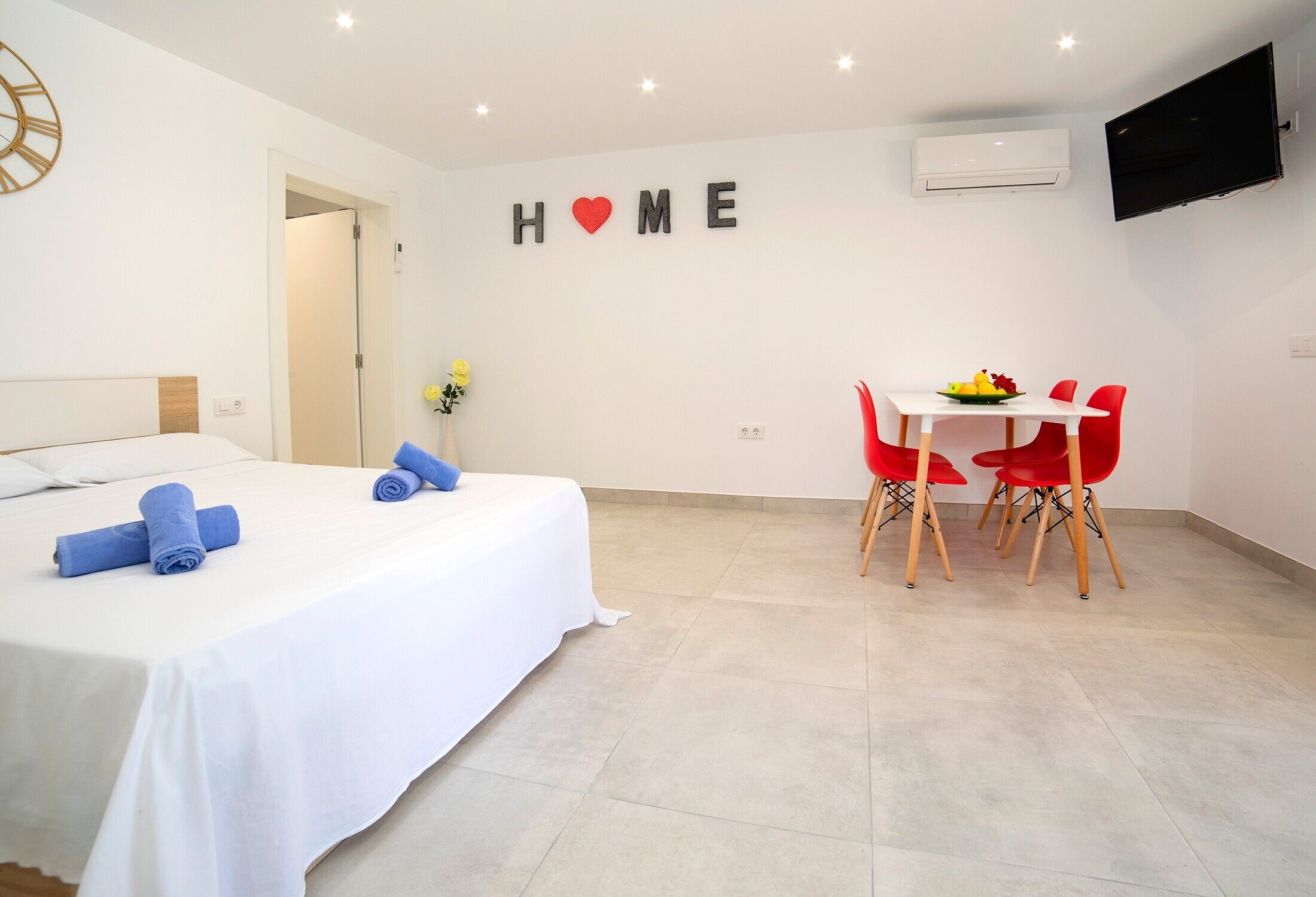 A hotel room with a bed, a dining table with chairs, a TV, and an air conditioning unit.
