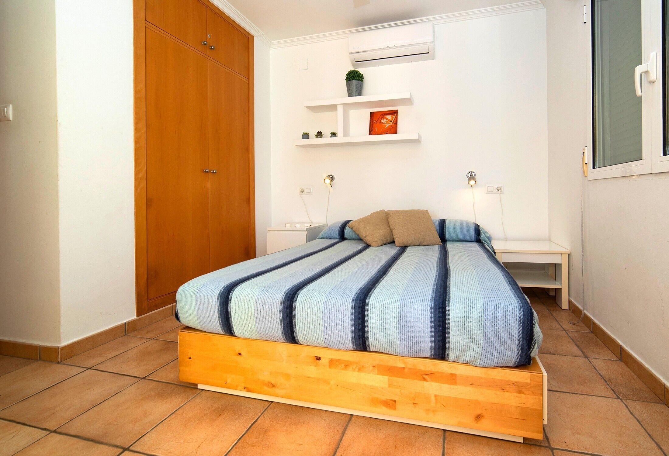 Un dormitorio con un cabecero de madera, una pared blanca, una pequeña estantería con adornos y una ventana.