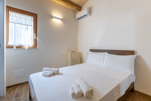A neatly made bed with white linens and towels, a window with sheer curtains, an air conditioning unit, and a wooden floor.