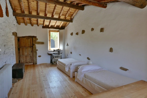 Une chambre rustique avec un plancher en bois, deux lits simples, un bureau avec une chaise, une fenêtre et un mur en pierre.