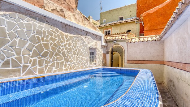 Ein Schwimmbecken mit Mosaikfliesen, einer Steinwand und einem Gebäude mit einem Flachdach im Hintergrund.