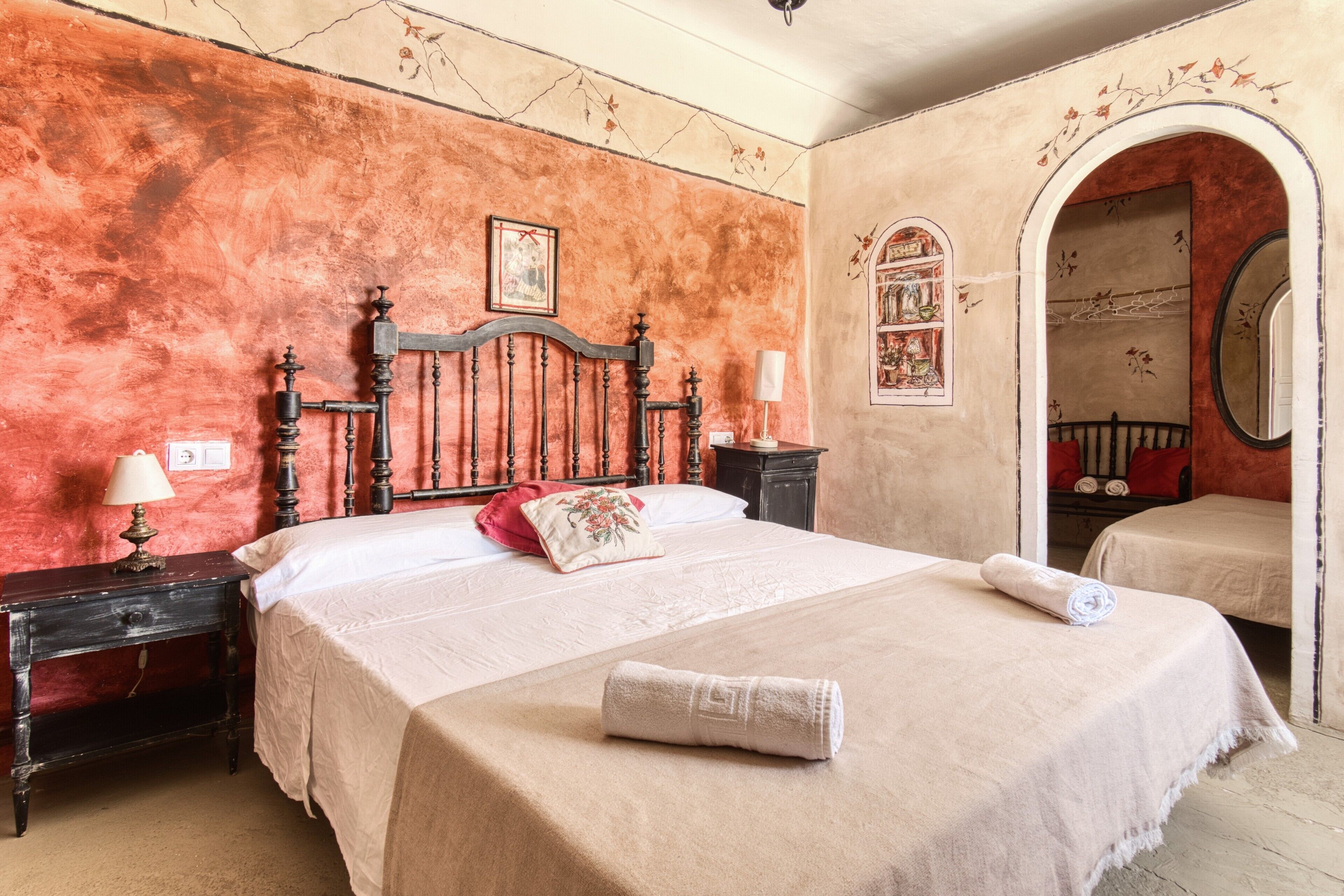 A bedroom with a large bed, a nightstand, a lamp, a red pillow, a rolled towel, and a mirror.