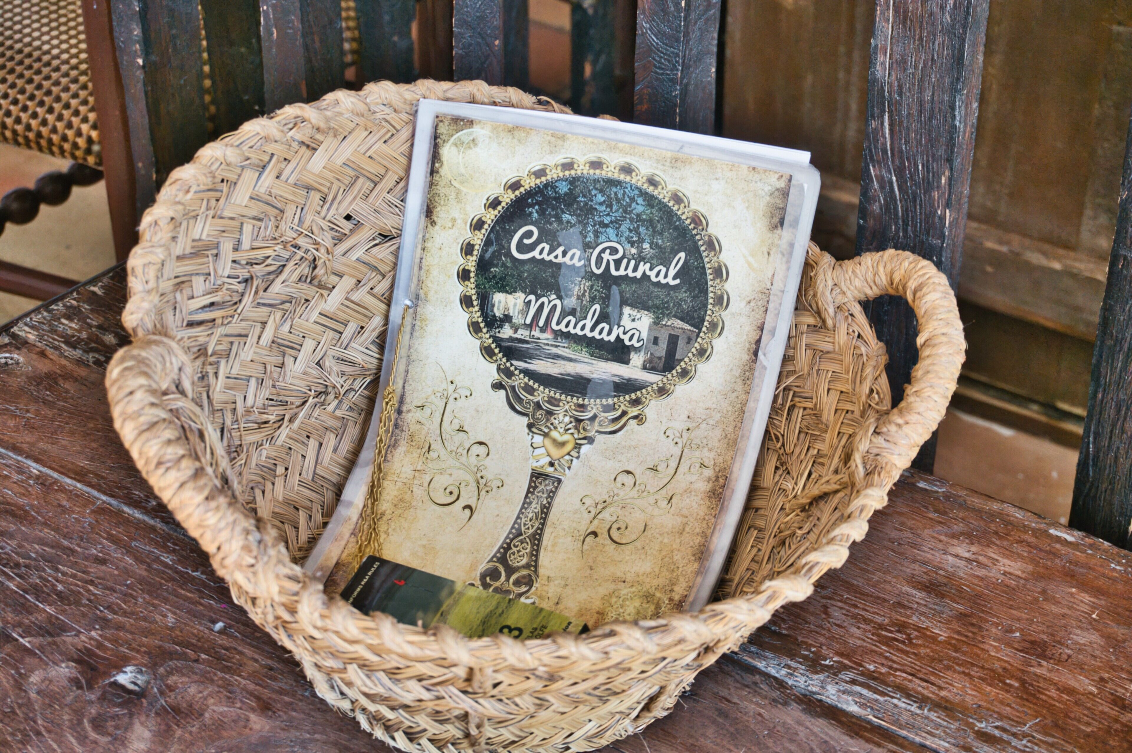 Een rieten mand met daarin een boek getiteld 'Casa Rural Madara' op een houten bank.