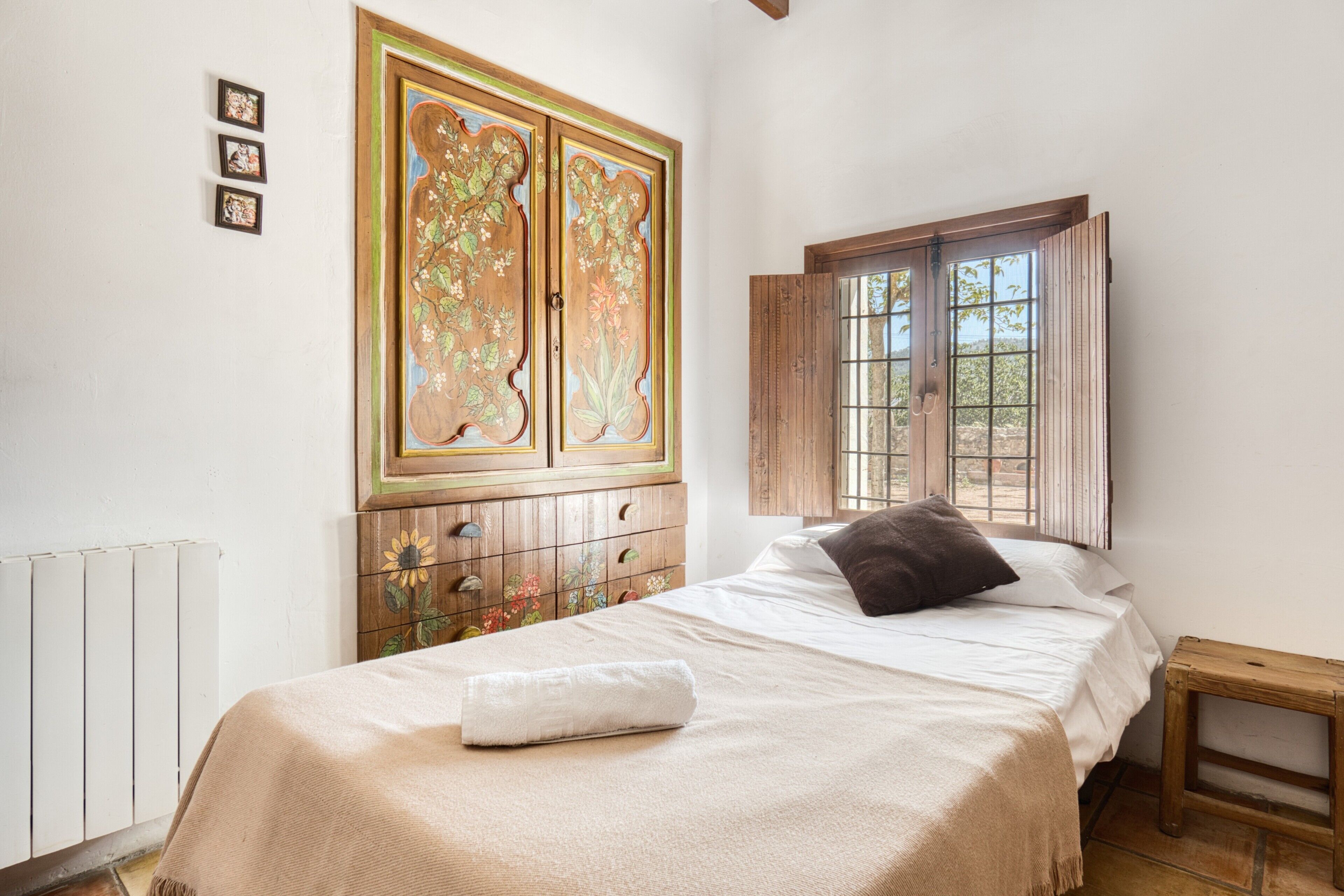 Un dormitorio con cama, un armario de madera con diseños florales, una ventana con contraventanas de madera y un radiador.