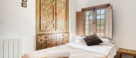 Un dormitorio con cama, un armario de madera con diseños florales, una ventana con contraventanas de madera y un radiador.