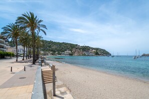 A coastal promenade with a sandy beach, palm trees, and a view of the sea with boats.