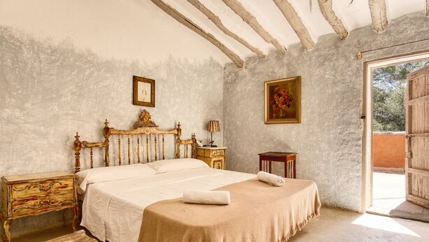 Une chambre avec un grand lit, une tête de lit en bois, des tables de chevet, une commode, une porte donnant sur l’extérieur et un tableau accroché au mur.