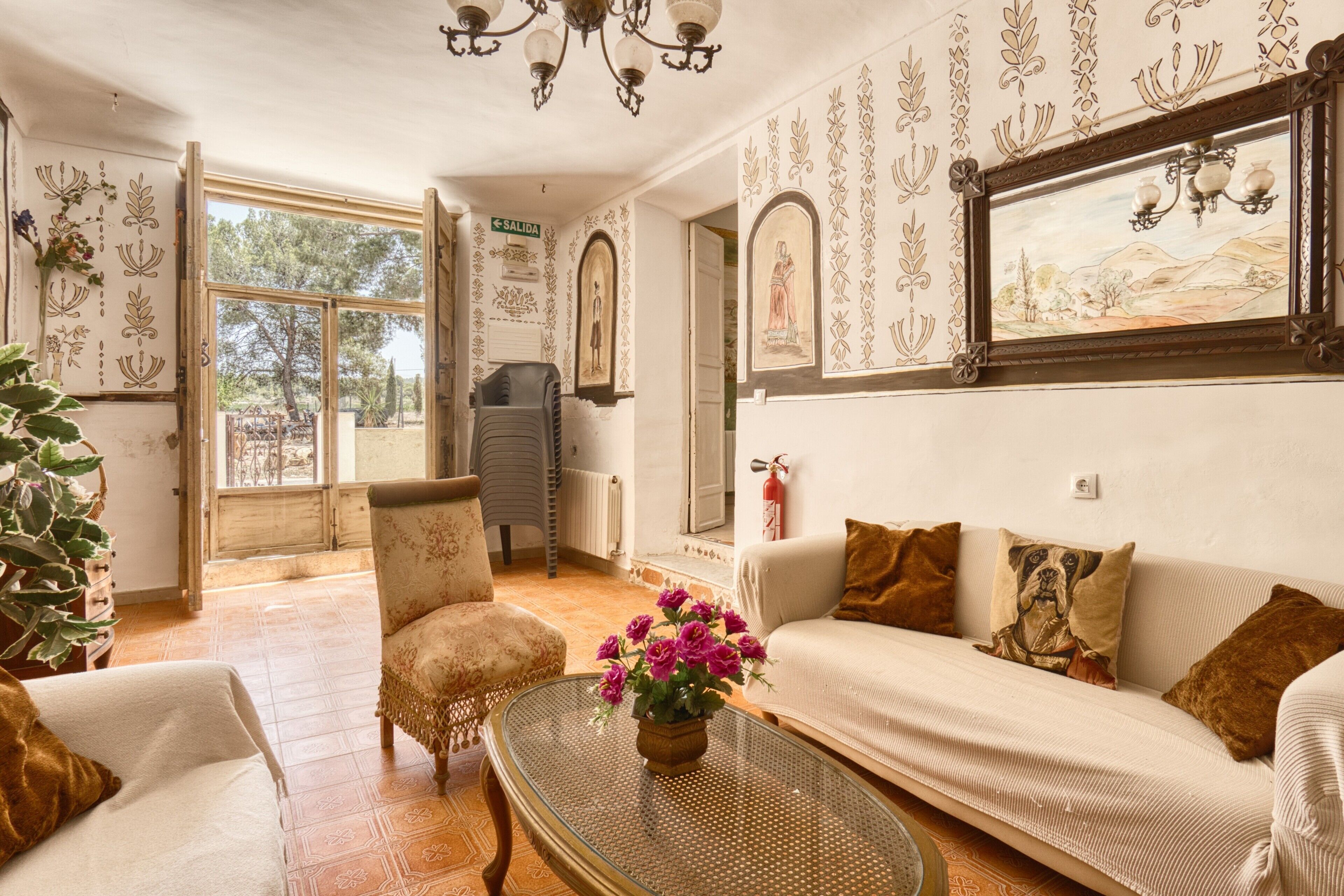 Habitación con sofá, sillón y una mesa con un florero. Se ve una ventana, una puerta y una pared decorada con cuadros.