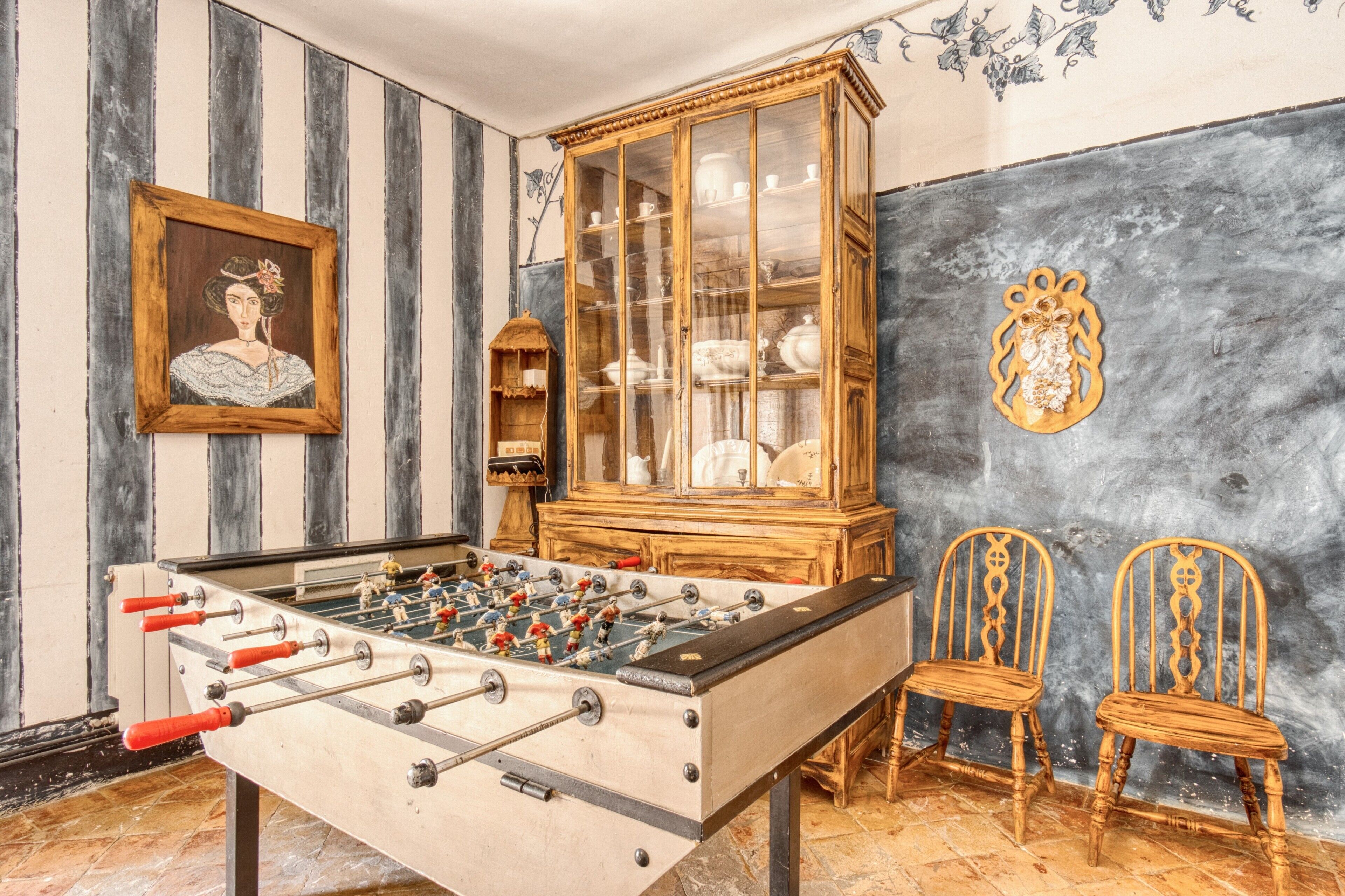 Una habitación con mesa de fútbol, un mueble de madera con puertas de cristal, dos sillas de madera y un cuadro en la pared.