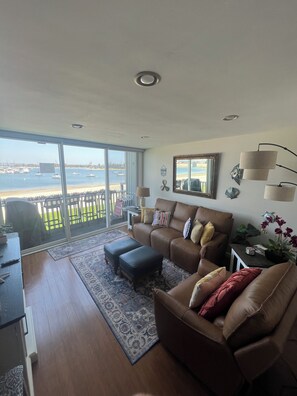 Uma sala de estar com uma janela grande com vista para uma marina, um sofá de couro marrom, uma mesa de centro e uma pequena mesa lateral com um abajur.