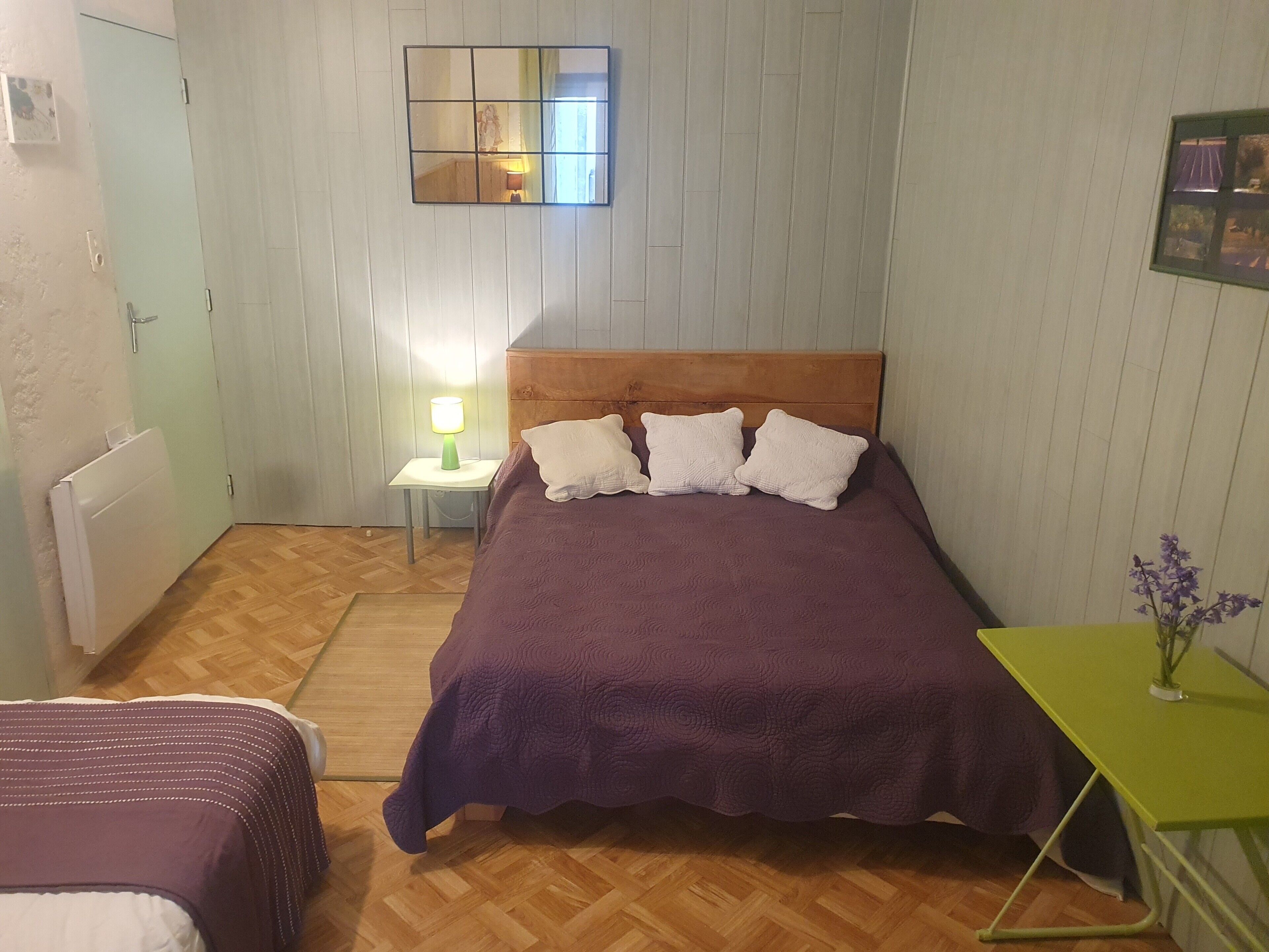 Une chambre avec un lit, une table de chevet avec une lampe, un radiateur et une petite table sur laquelle est posé un vase.