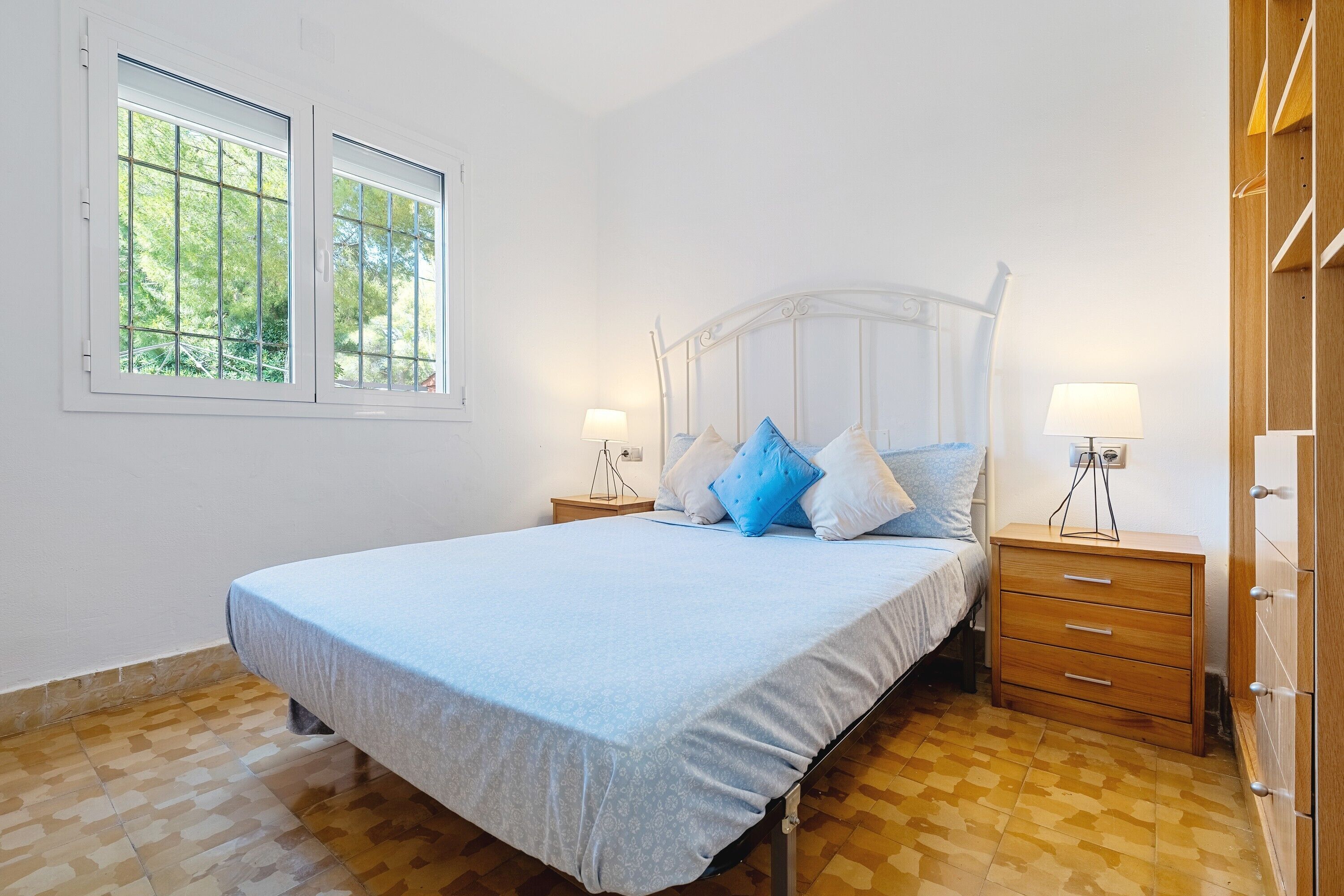 Une chambre à coucher avec un lit, deux tables de chevet, une commode en bois et une fenêtre donnant sur les arbres.