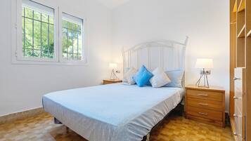 Une chambre à coucher avec un lit, deux tables de chevet, une commode en bois et une fenêtre donnant sur les arbres.