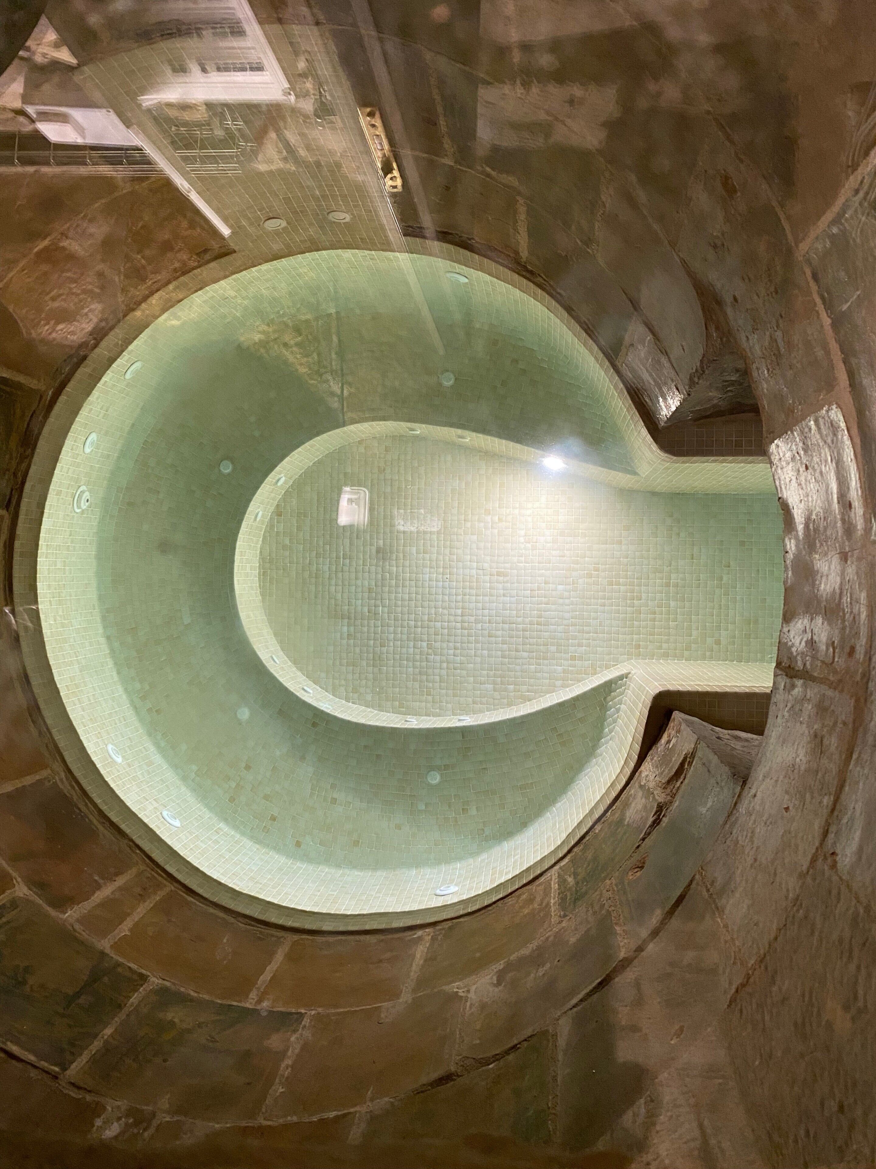 Una piscina circular con baldosas y una estructura central alargada, rodeada de muros de piedra.