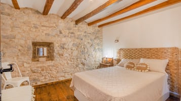 Une chambre avec un mur en pierre, des poutres apparentes, un lit doté d’une tête de lit en osier, une table de chevet et un miroir.