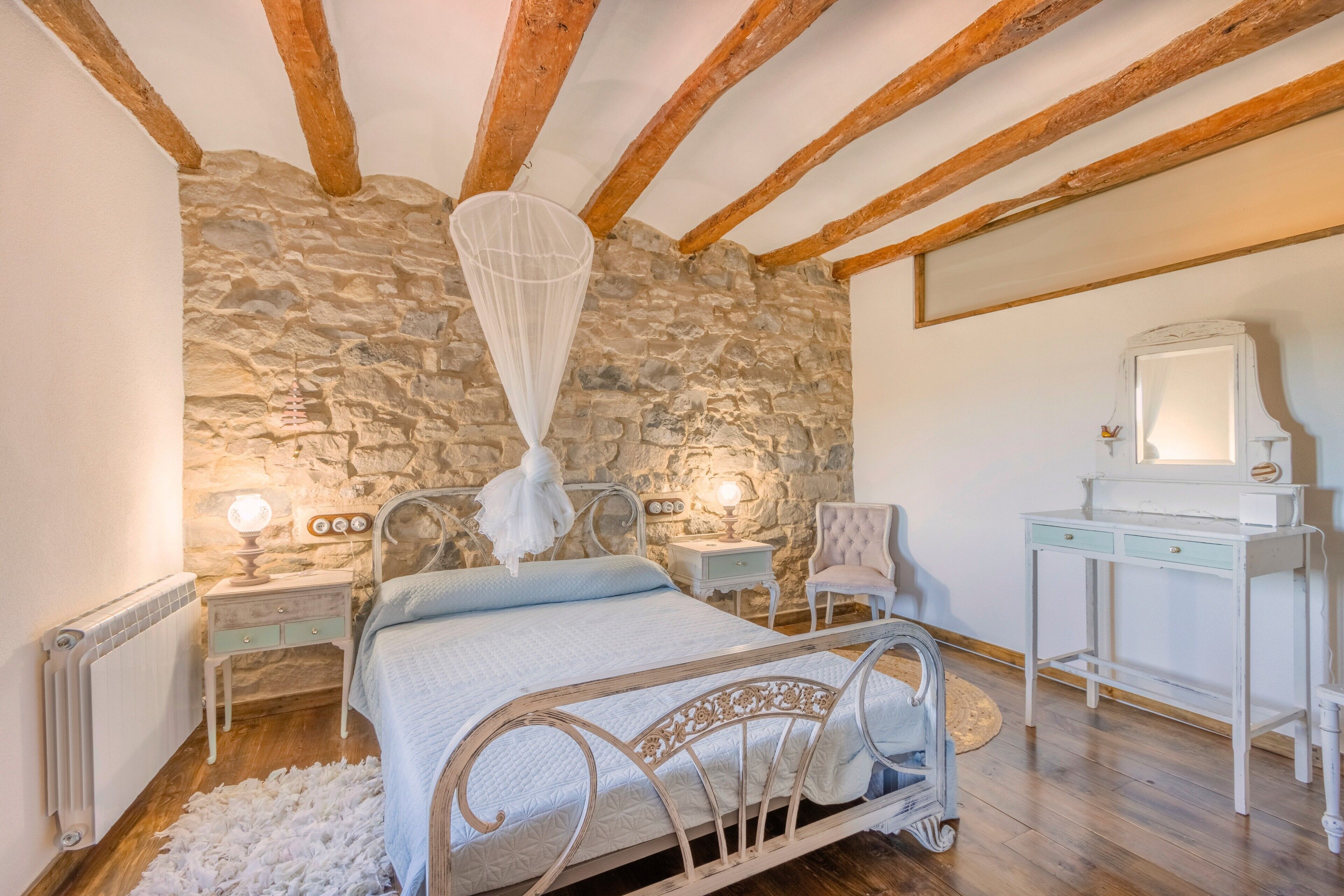 Une chambre avec un mur en pierre, un lit à baldaquin, une table de chevet avec une lampe, une chaise et un bureau.