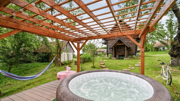 A hot tub under a wooden pergola in a garden with a hammock, wooden deck, and rustic buildings in the background. - 2 bedroom cozy home in Konjscina (Konjscina)