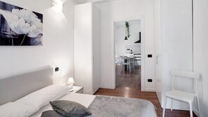 Ein modernes Schlafzimmer mit Bett, Nachttisch und einem Gemälde. Ein Durchgang führt zu einem Essbereich mit Tisch und Stühlen.