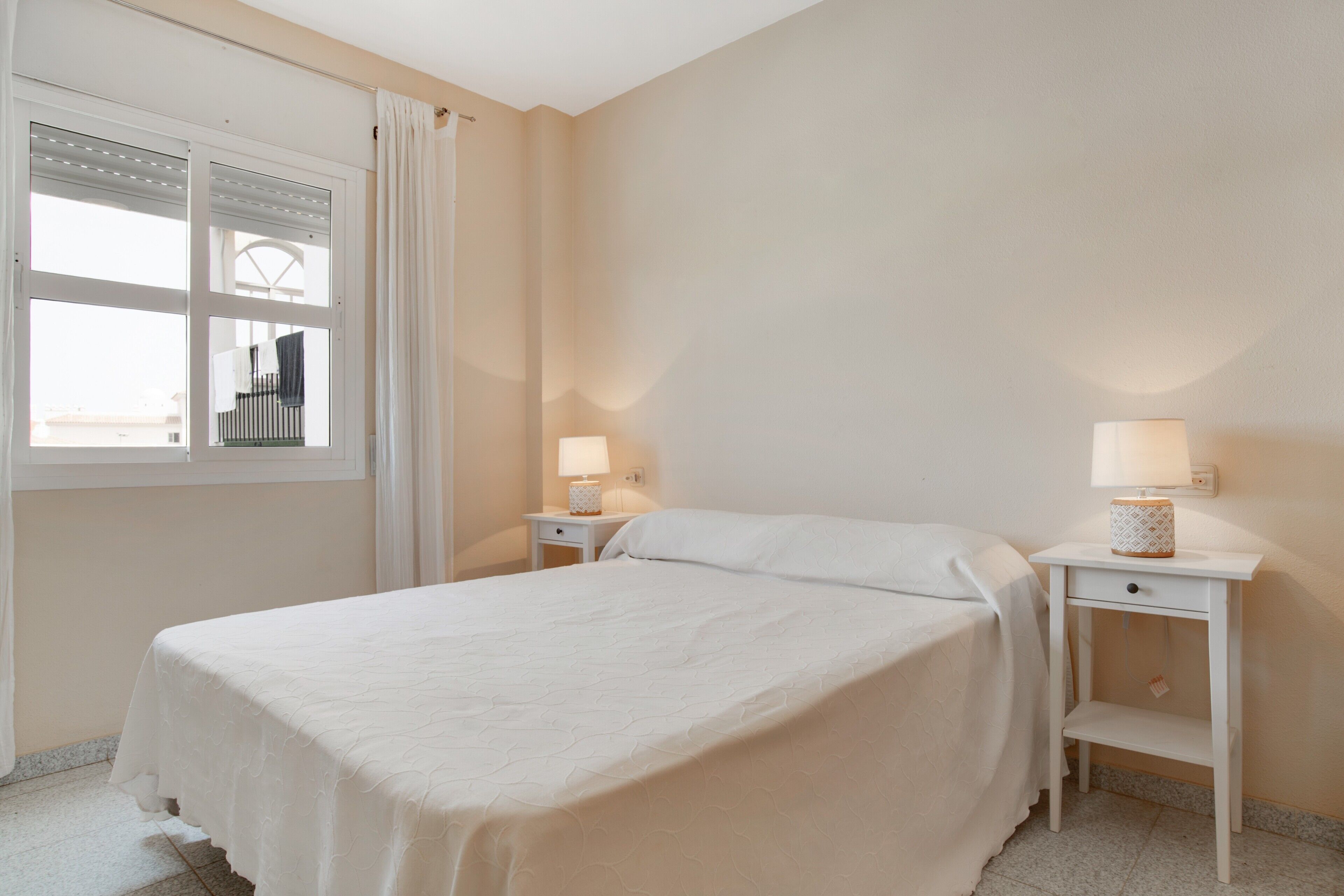 A bedroom with a bed, two bedside tables, a window with curtains, and a wall-mounted lamp.