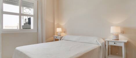 A bedroom with a bed, two bedside tables, a window with curtains, and a wall-mounted lamp.