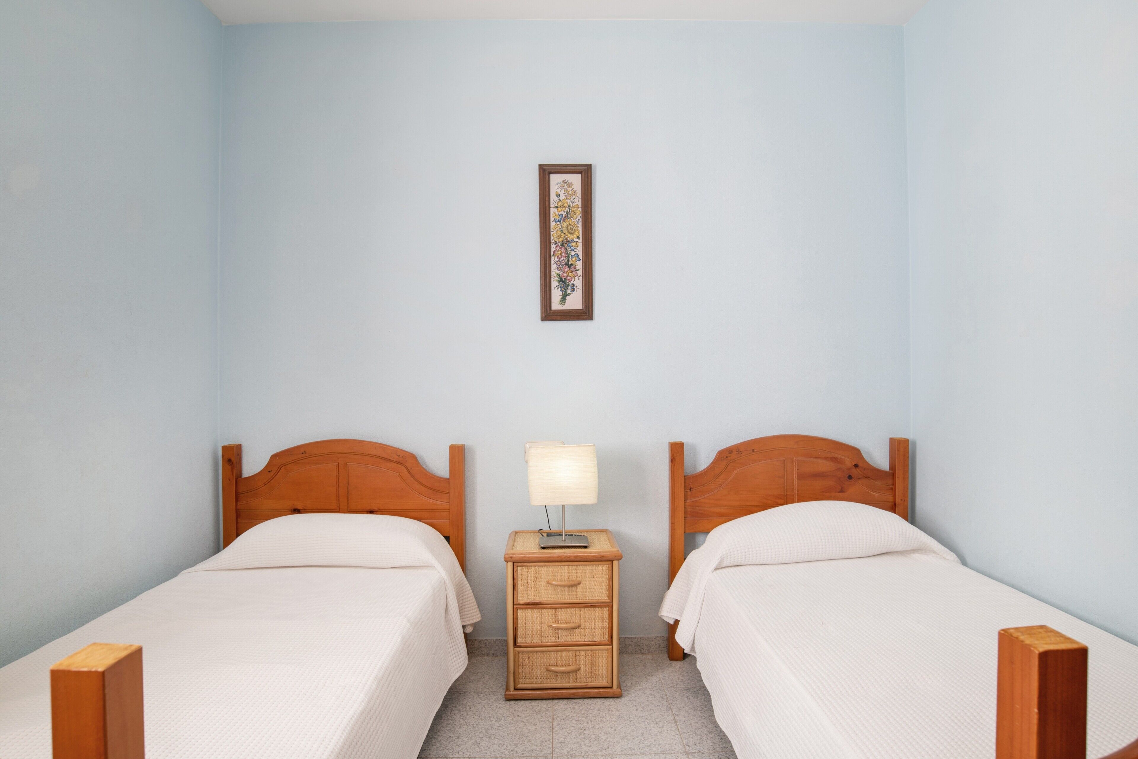A room with two wooden beds, a nightstand with a lamp, and a framed picture on the wall.