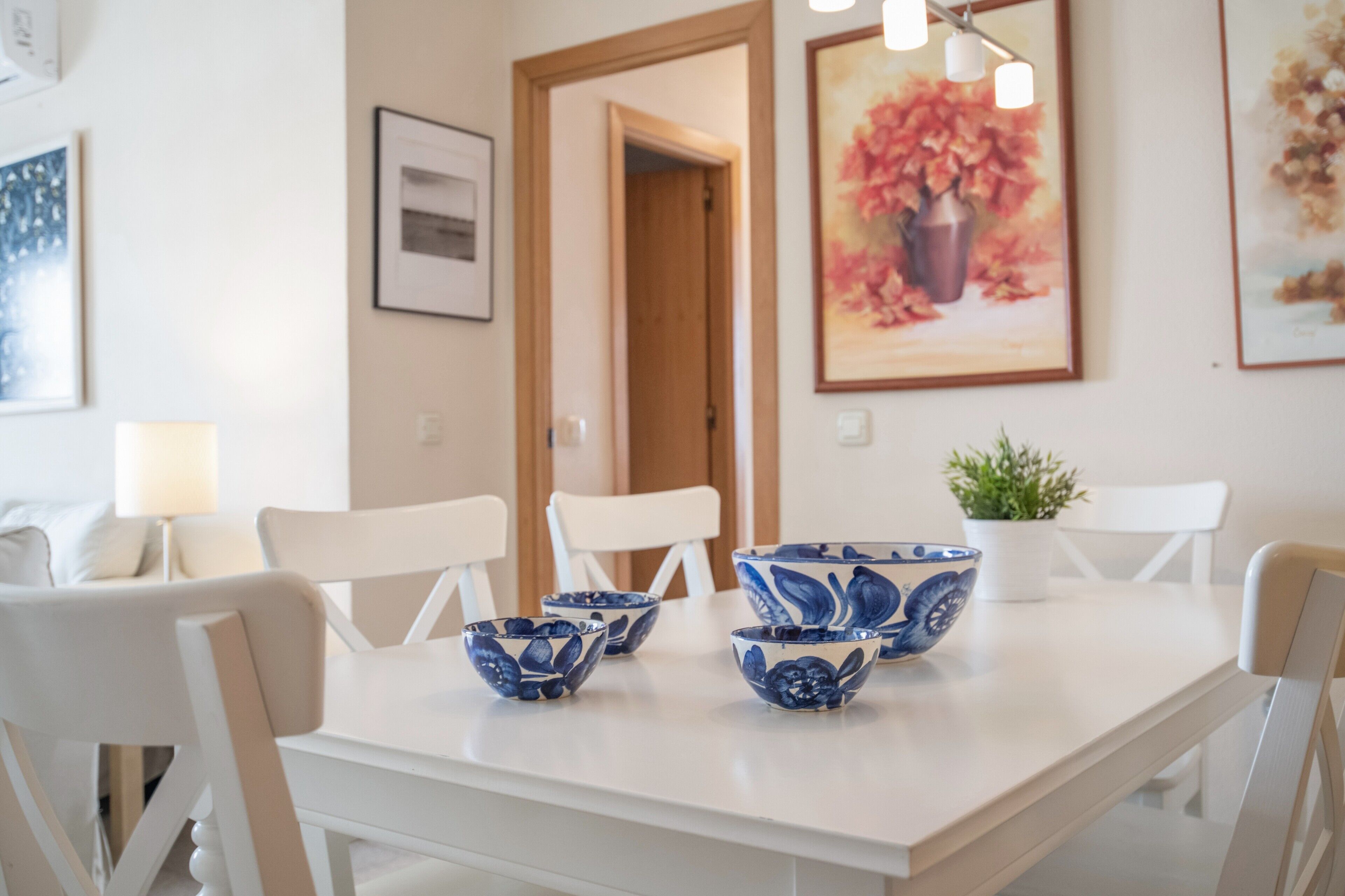 A dining area with a white table, blue and white patterned bowls, white chairs, a potted plant, and framed artwork on the walls.