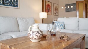 Una sala de estar con una mesa de centro de madera, sofá blanco, un jarrón decorativo, botella de vino y copas.