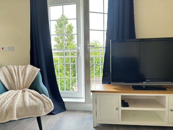 Une pièce avec une grande fenêtre, un téléviseur à écran plat posé sur un meuble télé en bois, un fauteuil moelleux et un meuble blanc contenant une télécommande.