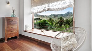 Une pièce comprenant une commode en bois, un fauteuil en osier avec des coussins blancs, et une grande fenêtre donnant sur un paysage verdoyant et montagneux.