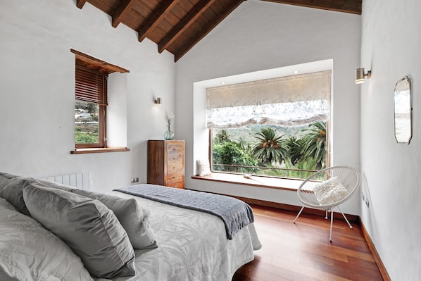 Une chambre à coucher avec un lit, une fenêtre donnant sur un paysage verdoyant, une chaise et une commode.
