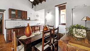 Une cuisine avec des meubles de rangement en bois, une table à manger, des chaises, un luminaire suspendu et une plante en pot.