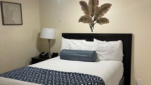 Una cama bien hecha con una colcha estampada, una planta en la pared, un ventilador de techo y una lámpara de noche.