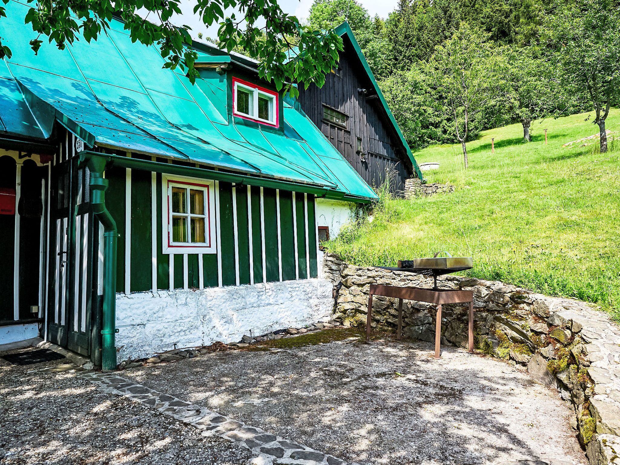 A traditional wooden house with a green roof and white trim, set in a rural landscape with a stone wall and a gravel path.