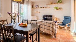 A dining area with a table, chairs, a fruit bowl, a TV, a ceiling fan, and shelves with decorative items. - Costabravaforrent Can Riera (l'Escala)