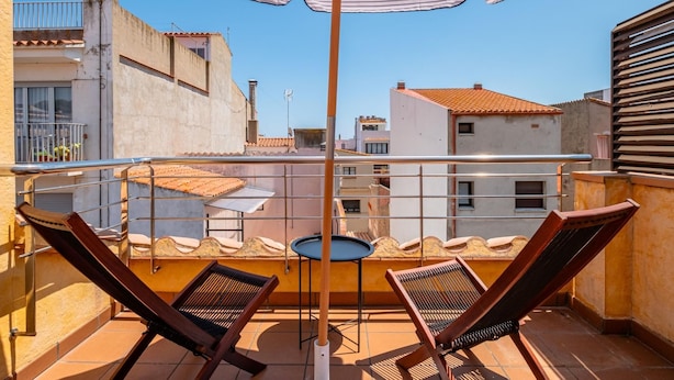 A balcony with two wooden chairs, a table, and a striped umbrella. - Costabravaforrent Can Riera (l'Escala)