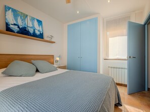 Een slaapkamer met een bed, kussens, een nachtkastje, een lamp, een schilderij, een kledingkast en een raam.
