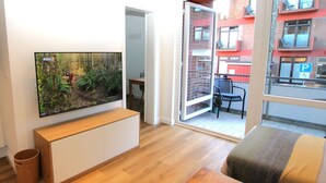 A modern living room with a flat-screen TV mounted on the wall, a wooden bench, a small table with coasters, and a sliding glass door leading to a balcony with a chair and a table. - Sandwall 60, Whg. 12, Huus in Lee - Ferienwohnung mit 3 - Zimmern auf ca. 50m² (Wyk auf Föhr)