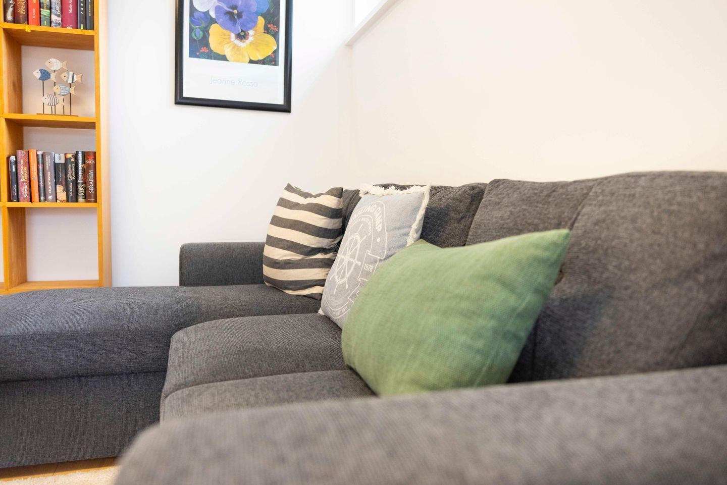 A grey sectional sofa with striped and green cushions, a bookshelf with books, and a framed picture of flowers on the wall.