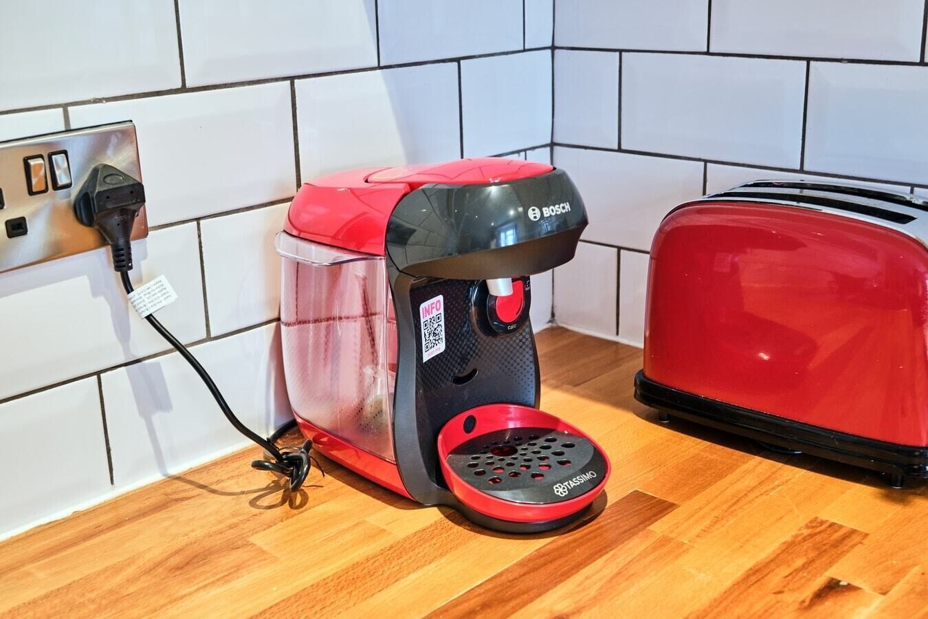 Een koffiezetapparaat met een rood en zwart design, aangesloten op een stopcontact in een betegelde muur.