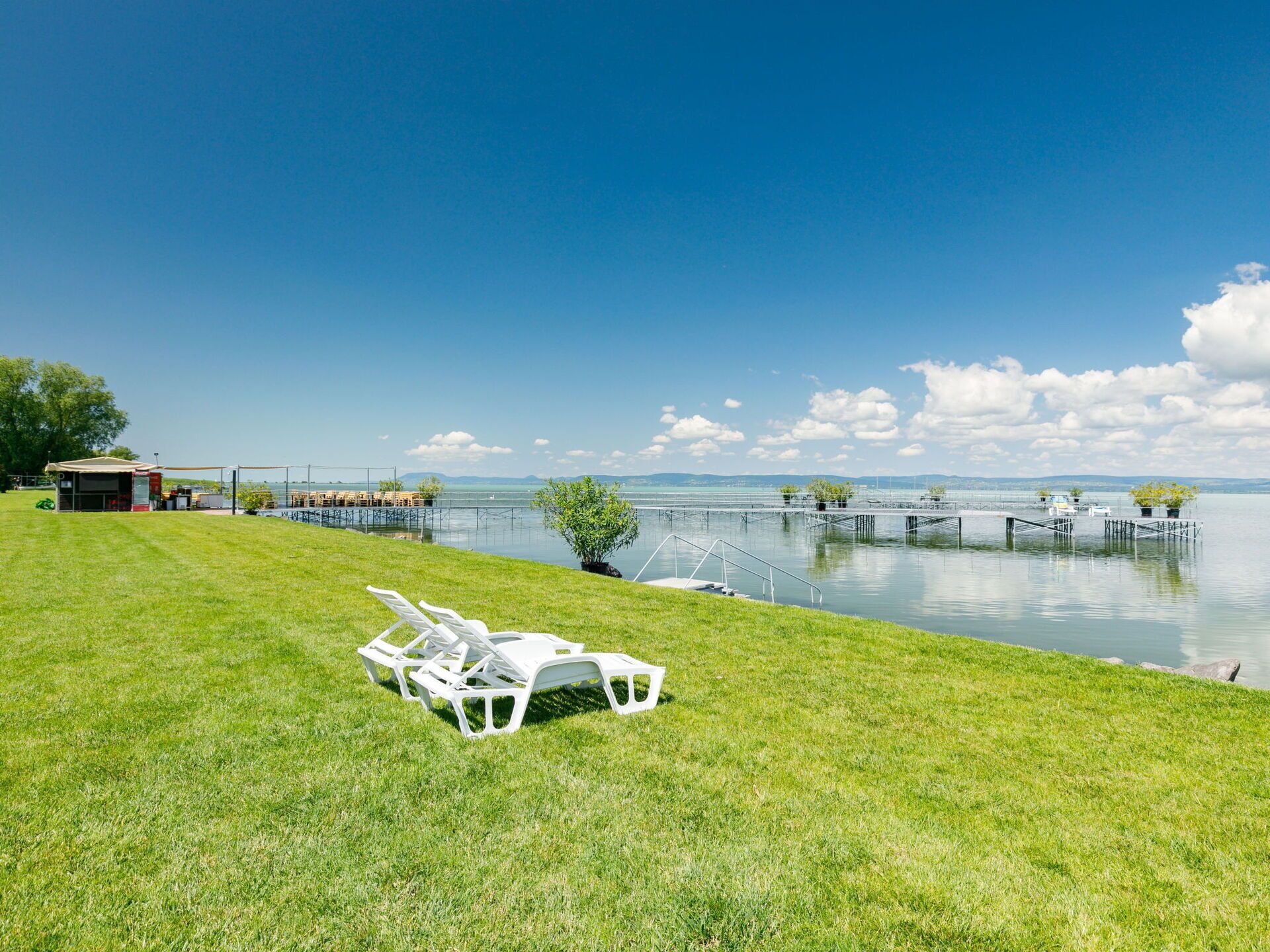 A grassy area with white lounge chairs and a small structure, likely a restroom, near a body of water with docks and boats.