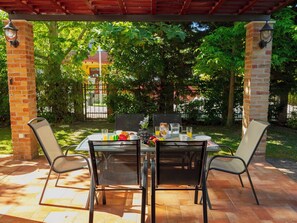 Une terrasse extérieure avec une table dressée pour deux personnes, entourée de piliers en briques, et une vue sur un quartier résidentiel avec des arbres et une porte au fond.