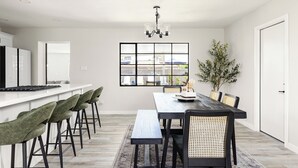 Une cuisine moderne avec un comptoir blanc, des tabourets de bar verts, une table à manger en bois avec des chaises noires et une grande fenêtre donnant sur l’extérieur.