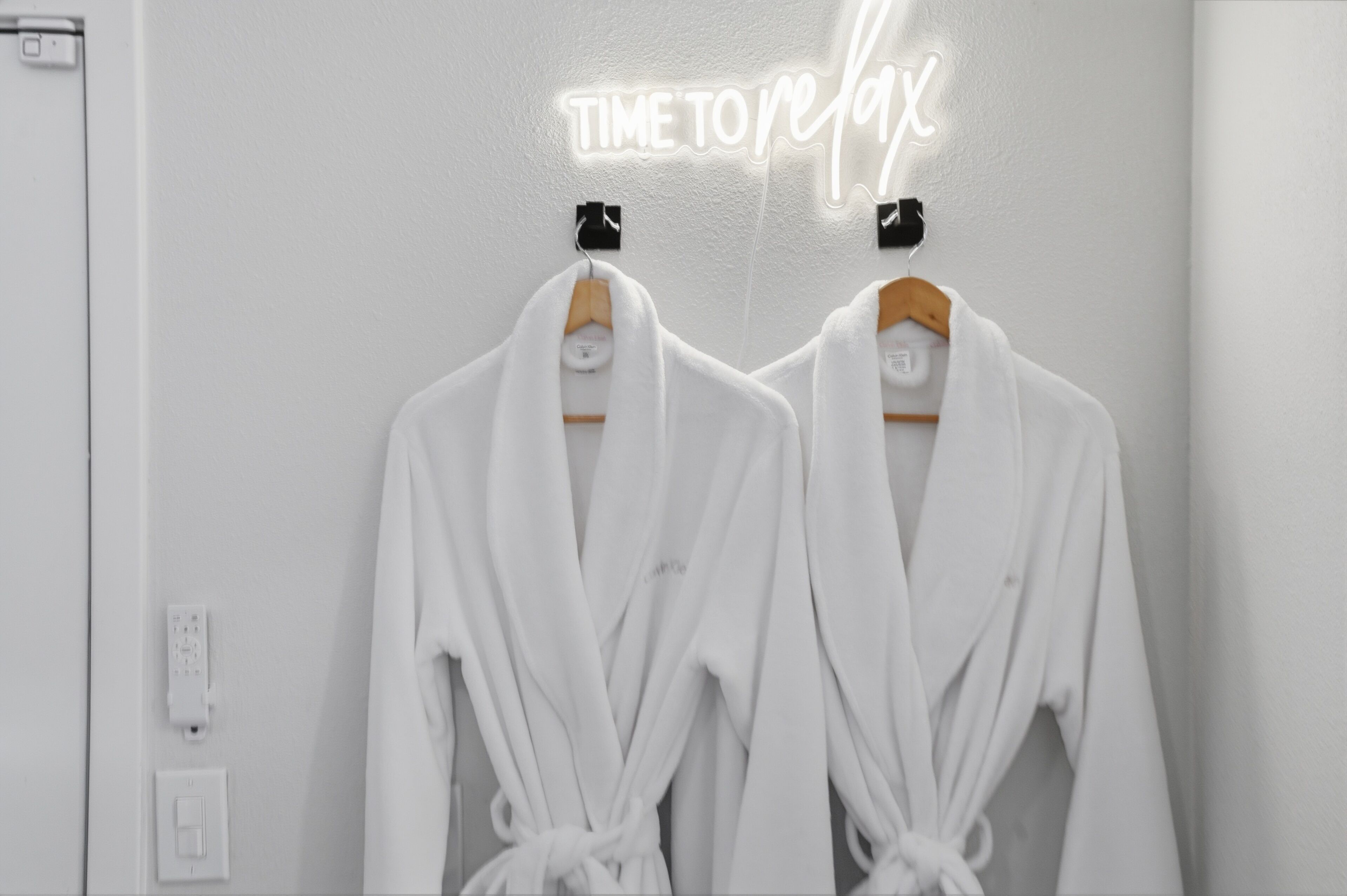 Dos batas blancas colgadas en perchas de madera frente a una pared con un letrero de neón que dice 'TIME TO REX'.