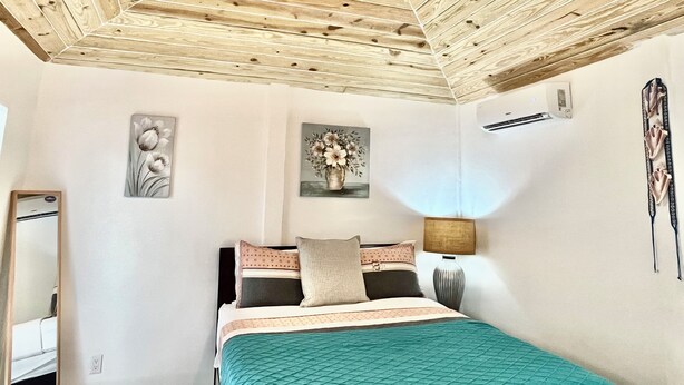 Ein Schlafzimmer mit Holzboden, einem Bett mit vielen Kissen, einem Nachttisch mit Lampe und Wandbildern.