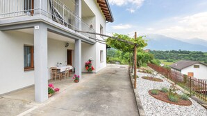 Un bâtiment à deux étages doté d’un balcon, d’un coin repas avec une table et des chaises, et d’un jardin agrémenté de plantes et d’une tonnelle.