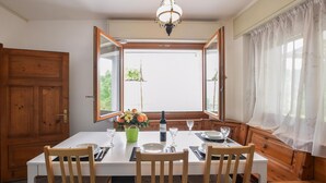 Une salle à manger avec une table dressée pour quatre personnes, un lustre et une vue dégagée sur l’extérieur grâce à de grandes fenêtres ouvertes.