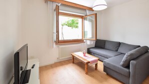 Un salon avec un canapé gris, une table en bois recouverte d’une nappe rose, un téléviseur posé sur un meuble et une fenêtre donnant sur un paysage verdoyant.