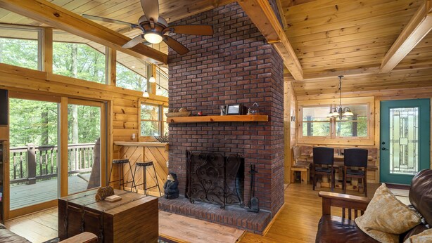 A cozy cabin interior with a fireplace, wooden furniture, and large windows overlooking a deck. - Cabin in the mountains with hot tub! (Ellijay)