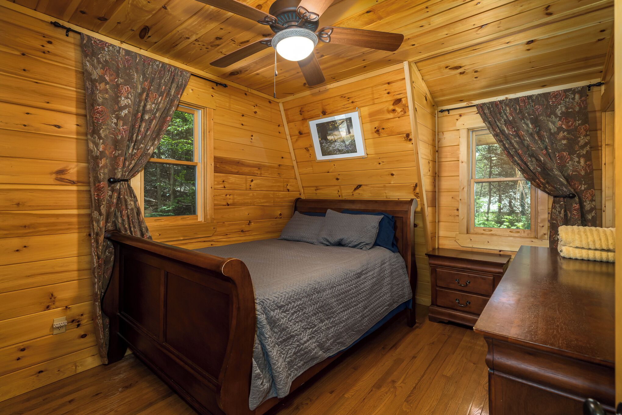 Un dormitorio de cabaña de madera con una cama, una mesita de noche, un aparador, un ventilador de techo y dos ventanas con cortinas.