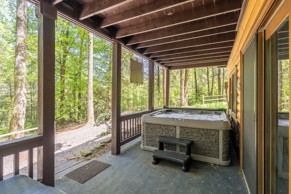 A covered outdoor hot tub area with a stone exterior, wooden deck, and a view of a forested area. - Cabin in the mountains with hot tub! (Ellijay)